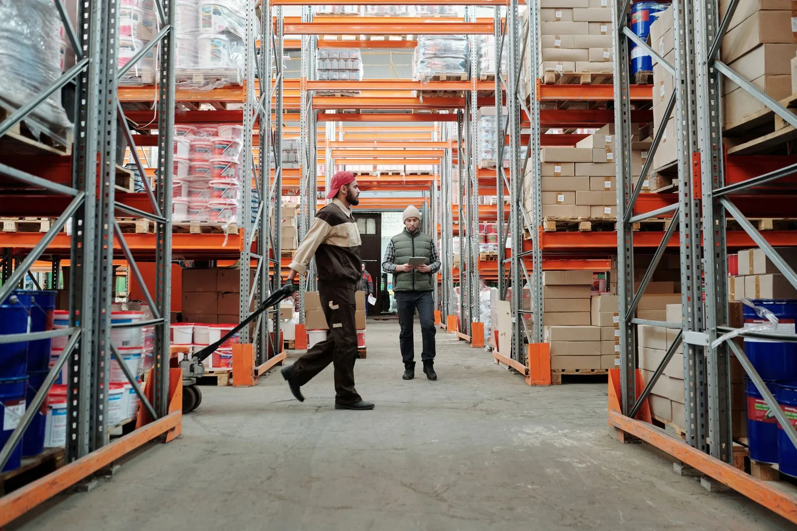 Two warehouse workers walking down a stocked orange-racking aisle with a pallet jack