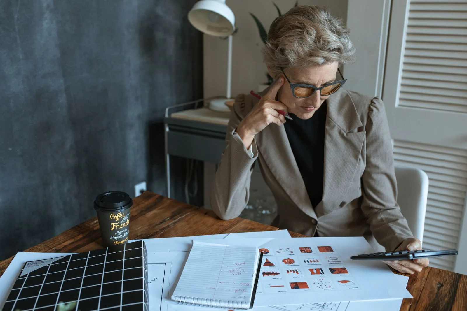 Senior woman with glasses studying printed documents and charts at a wooden desk
