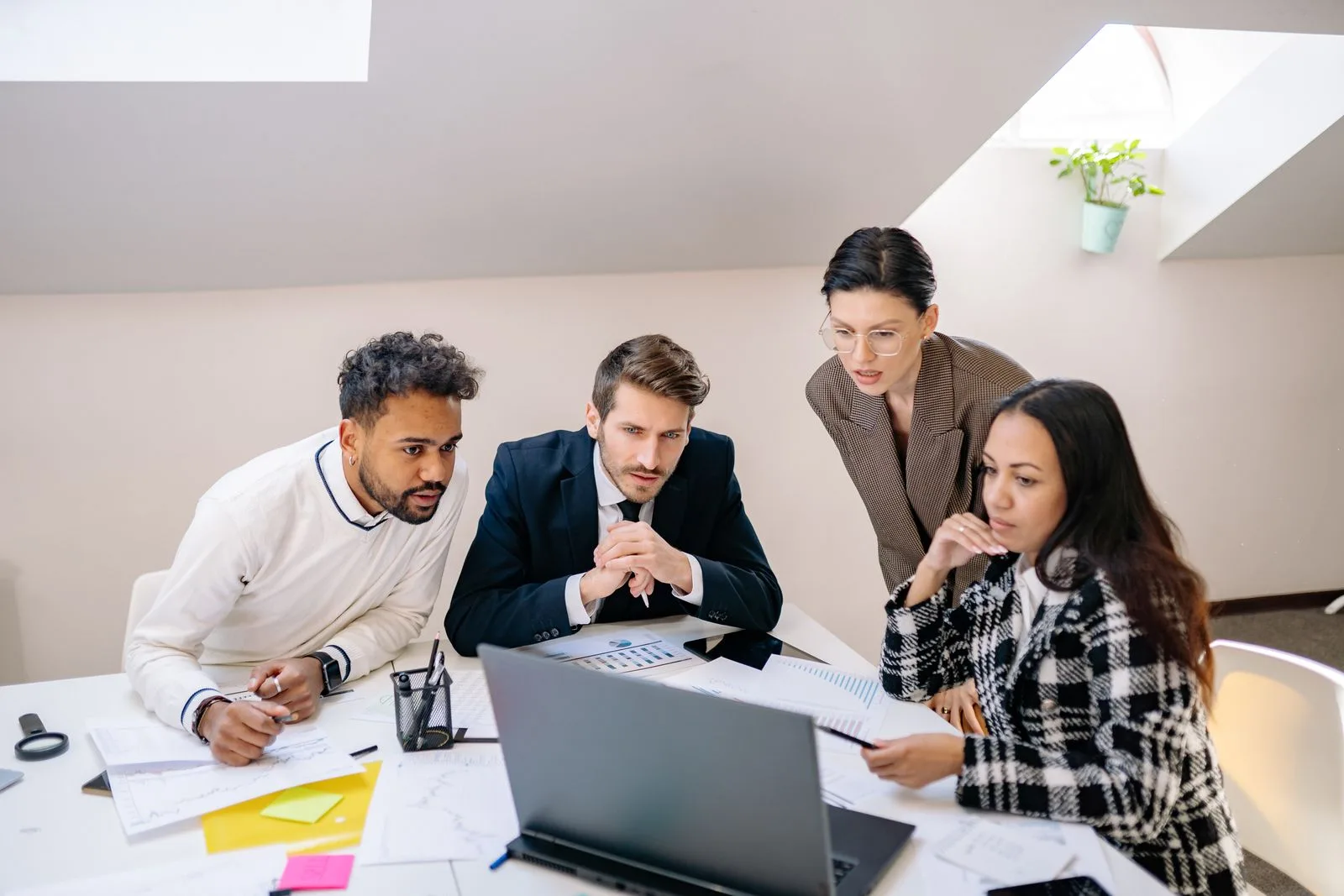 Four diverse business people gathered around a laptop reviewing printed reports in a bright office