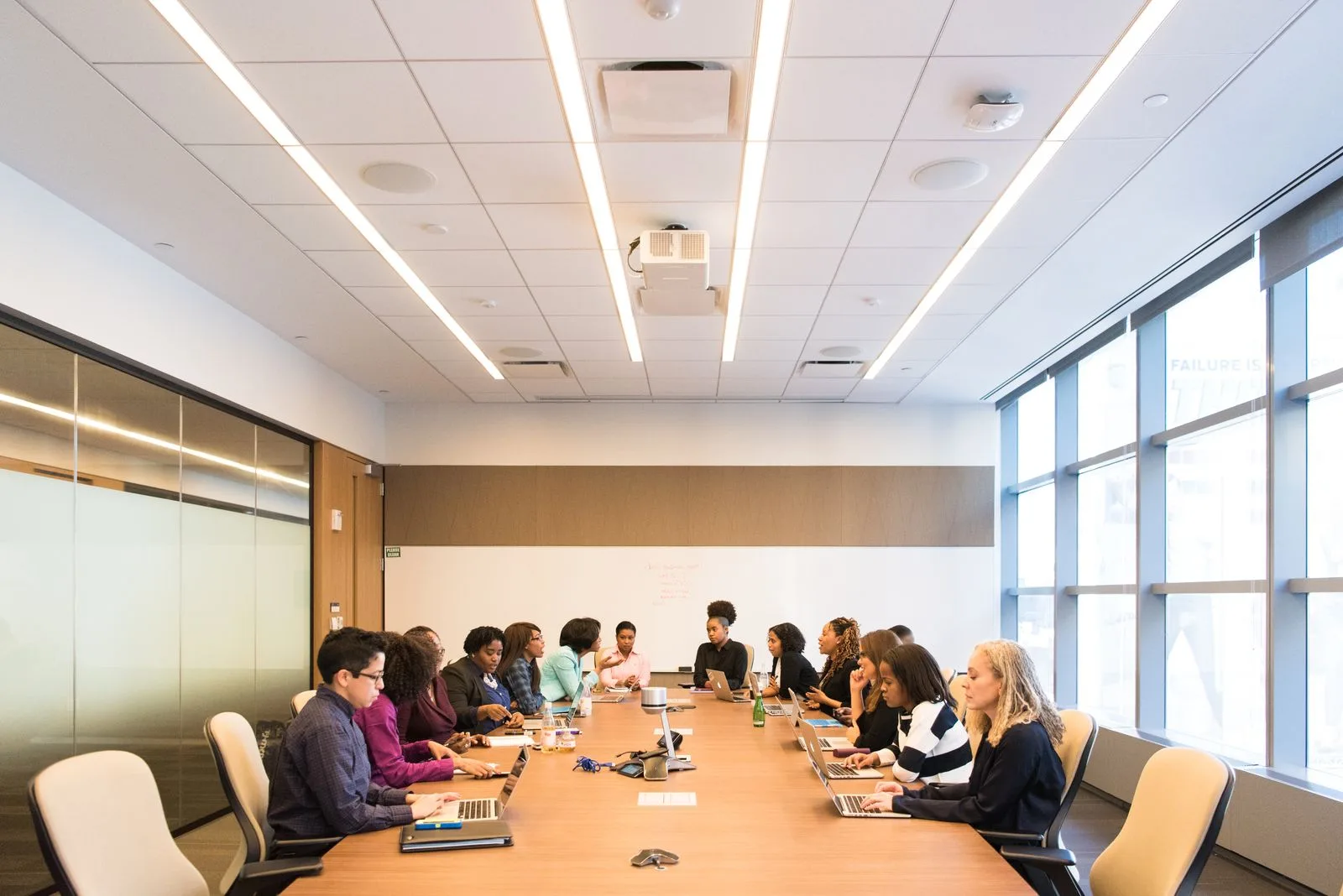 Large boardroom meeting with a long table of professionals seated with laptops in front of tall windows