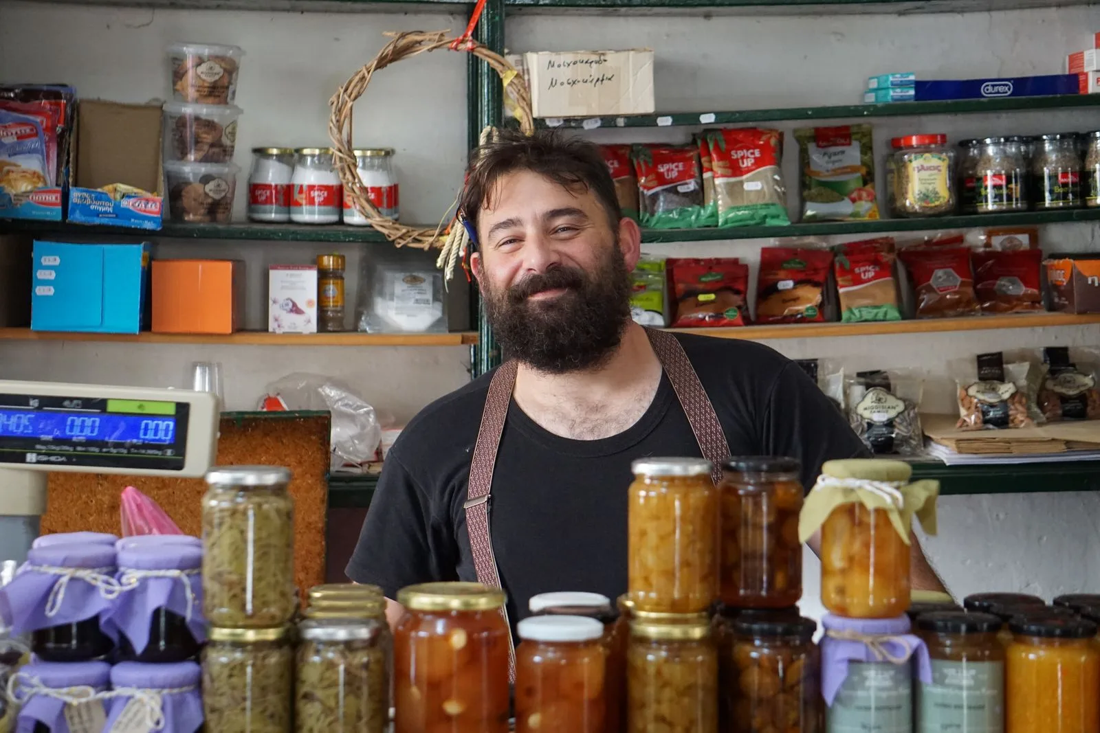 Small shopkeeper with beard and apron smiling behind a counter stacked with jars of preserves and pantry goods