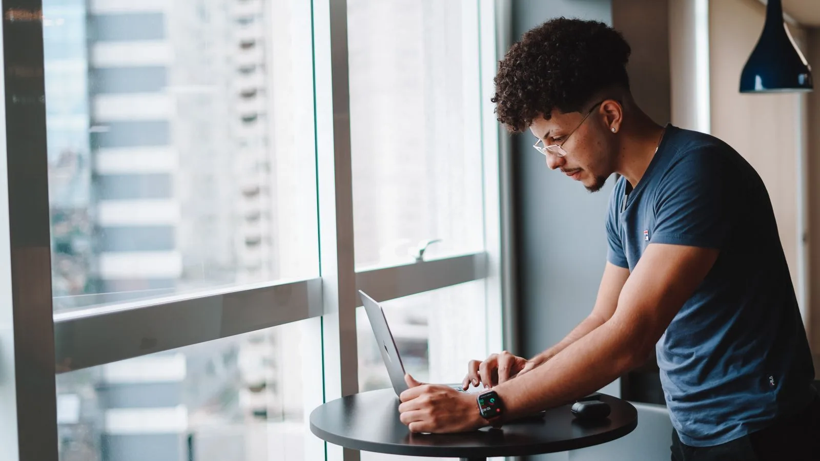 Young man in a blue t-shirt standing at a high table typing on a laptop by a floor-to-ceiling city window