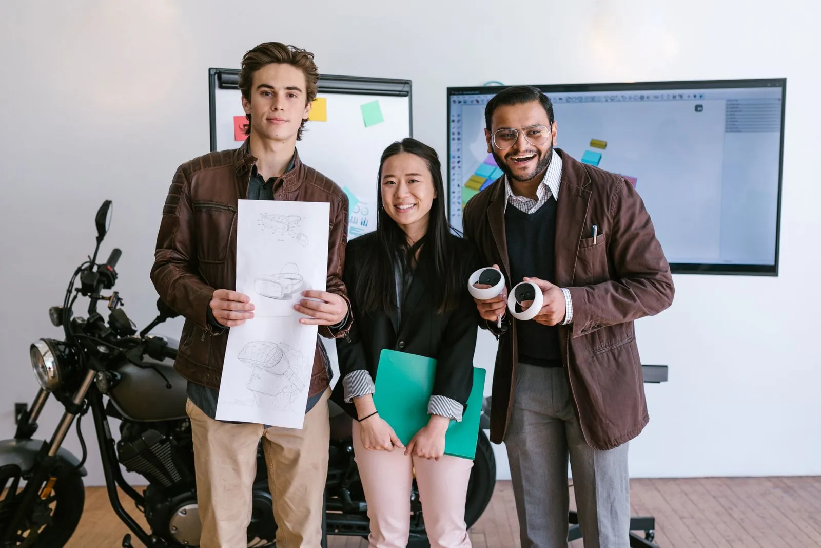 Three smiling startup founders standing together with a product sketch, VR controllers, and a design monitor behind them