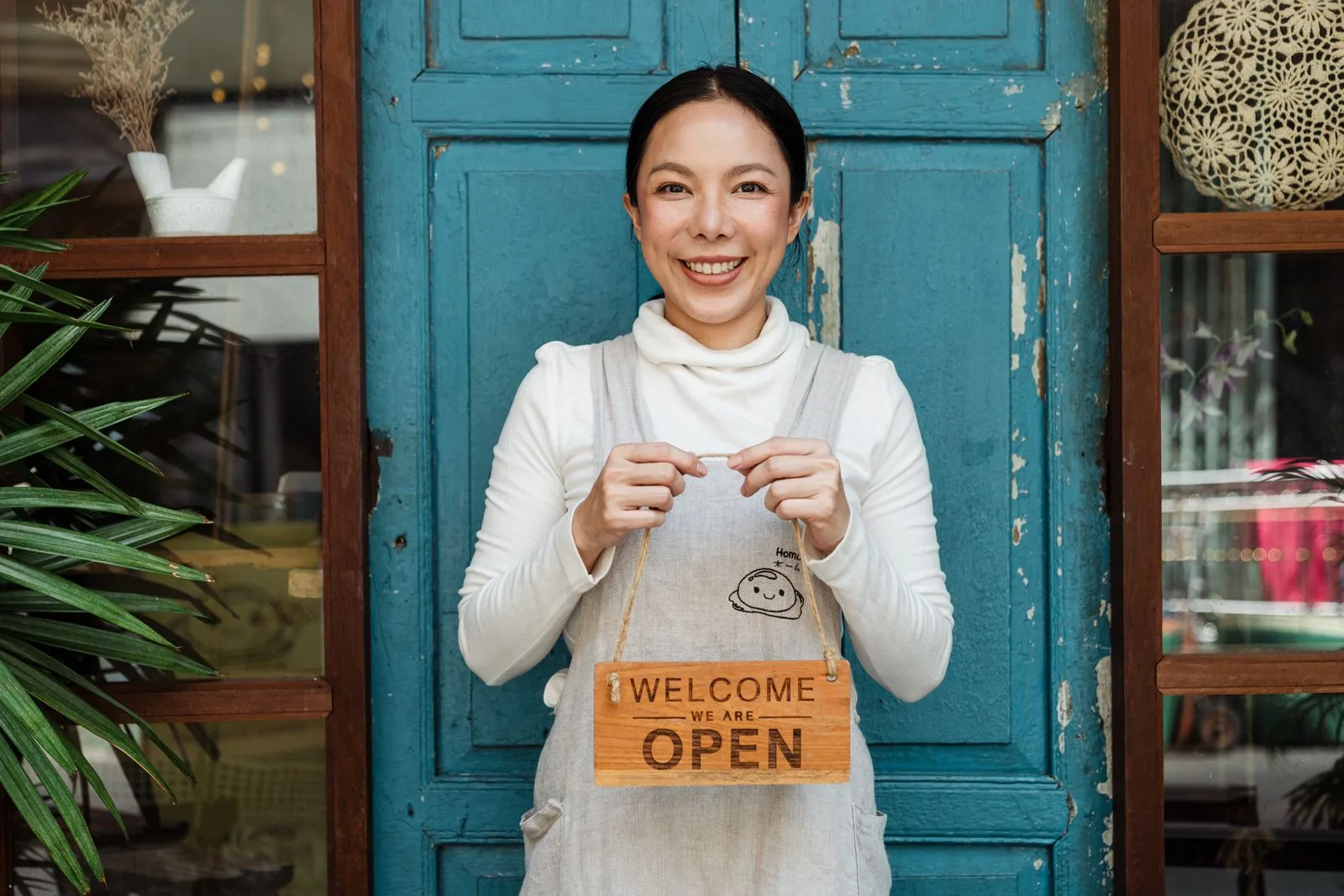 Small business owner smiling in front of a blue shop door holding a wooden Welcome We Are Open sign